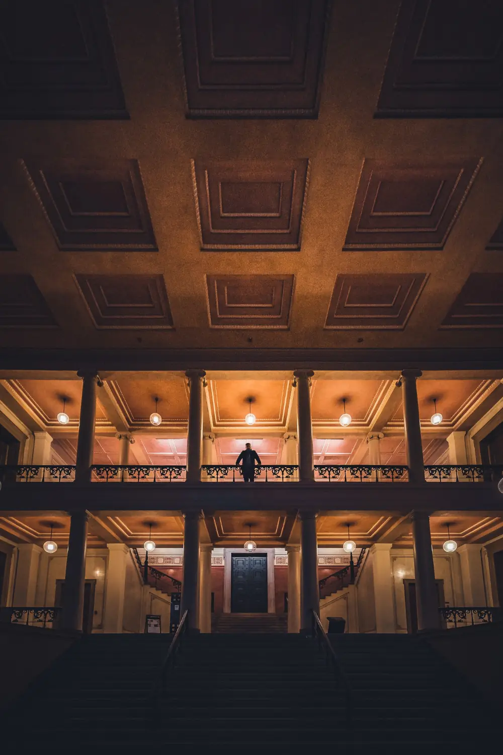 A person stands on a balcony inside a large building with tall columns, stairs, and warm lighting. The image represents Dot&’s Executive Search service for finding senior leaders and executive talent.